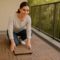 A young woman with a smile and open eyes installing interlocking WPC deck tiles on a balcony, surrounded by natural light and greenery in the background.
